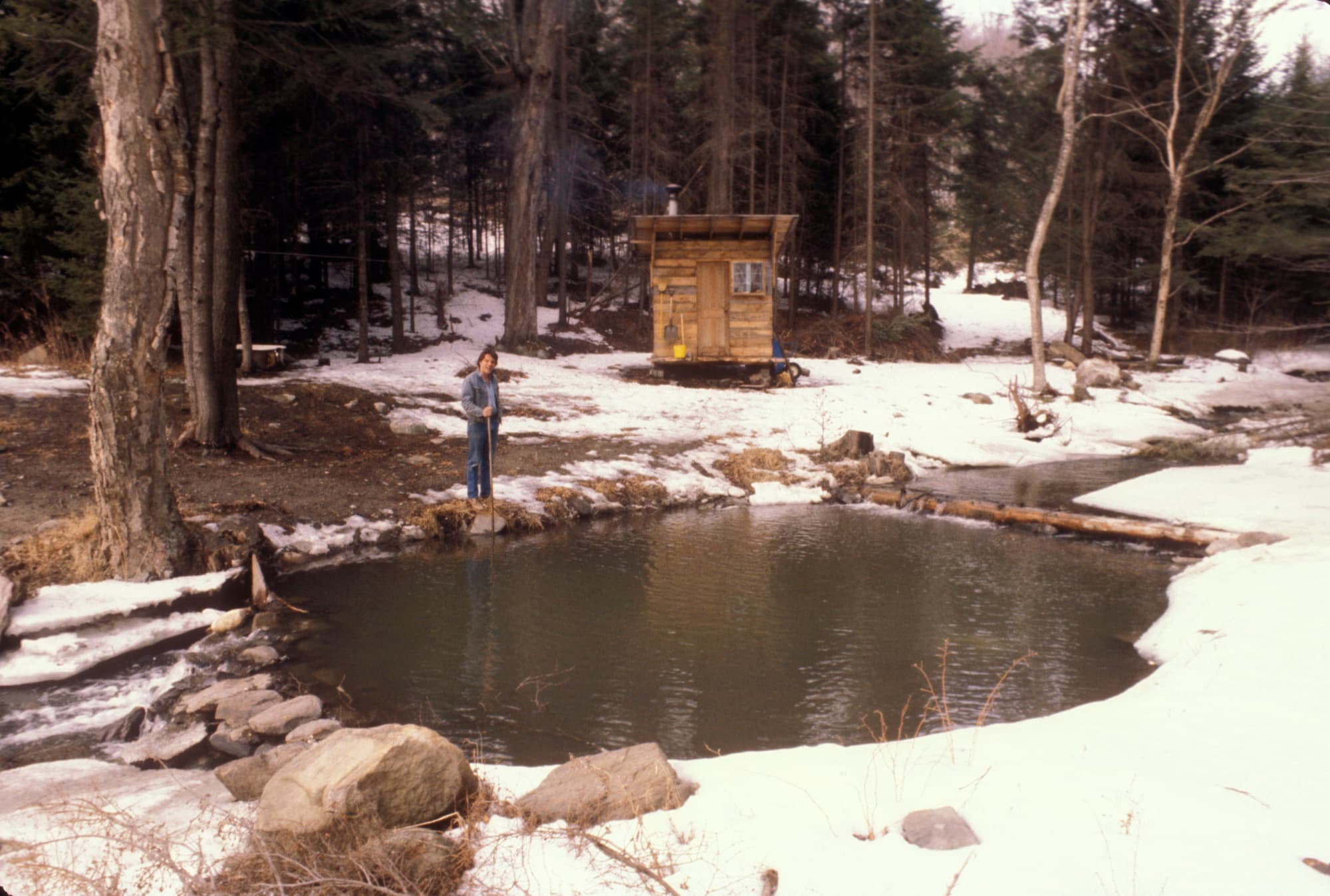 Blake Traendly in a denim jacket standing at the edge of the pond in late winter, snow on the banks, the small wooden sauna he built behind him with smoke rising from its stovepipe