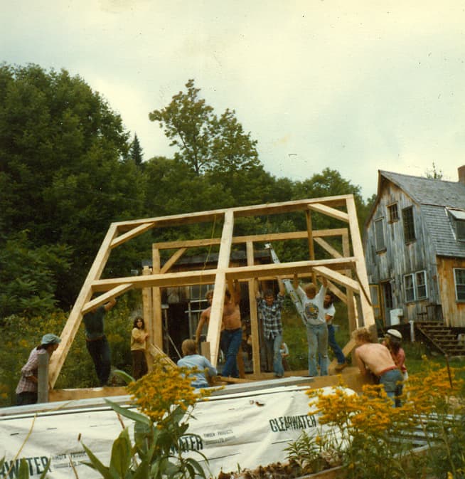 Friends in the early 1970s pushing the shop's first timber bent upright by hand, the old farmhouse behind them and goldenrod in the foreground
