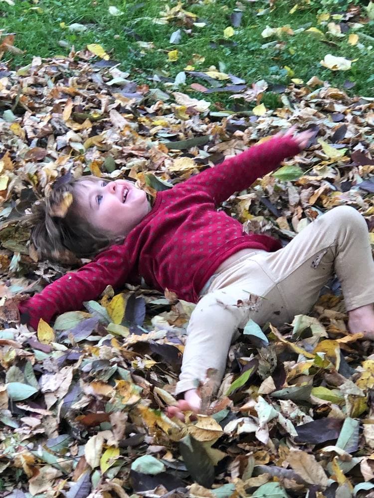 A kid grinning flat on her back in a pile of fall leaves