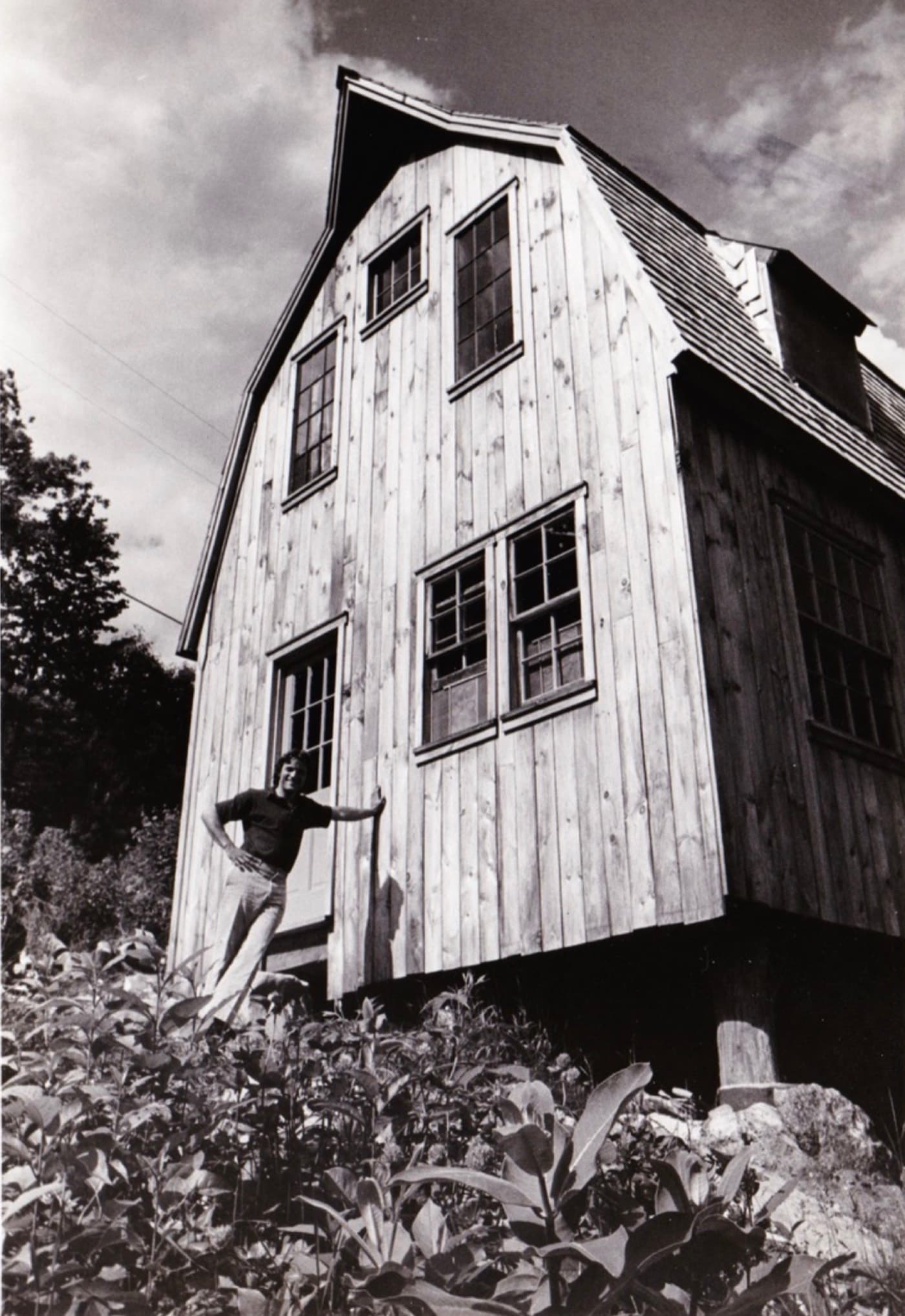 Black and white photograph of a young Blake Traendly standing proudly beside the tall gambrel-roofed house he built