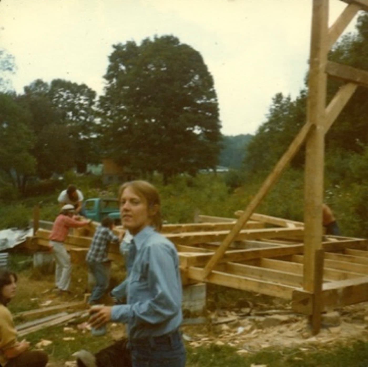 A faded color photograph from the early 1970s of friends raising the shop's timber frame in a Vermont field, a green pickup truck behind them