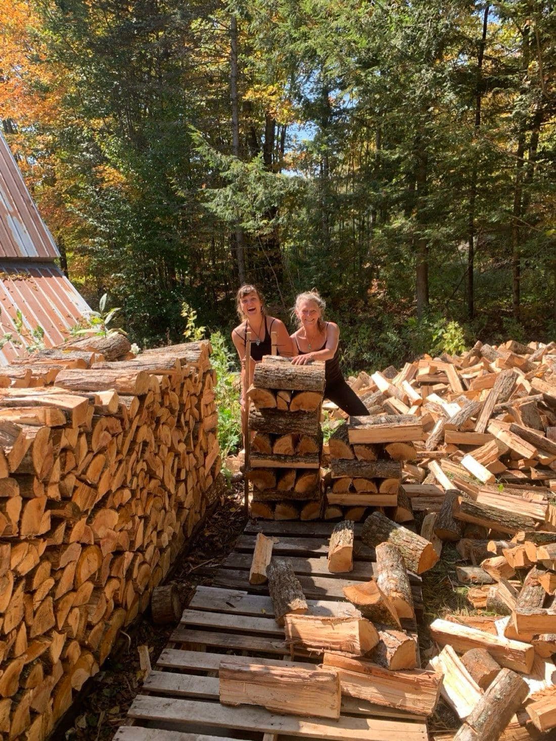 Two family members grinning behind an enormous woodpile on a sunny fall day
