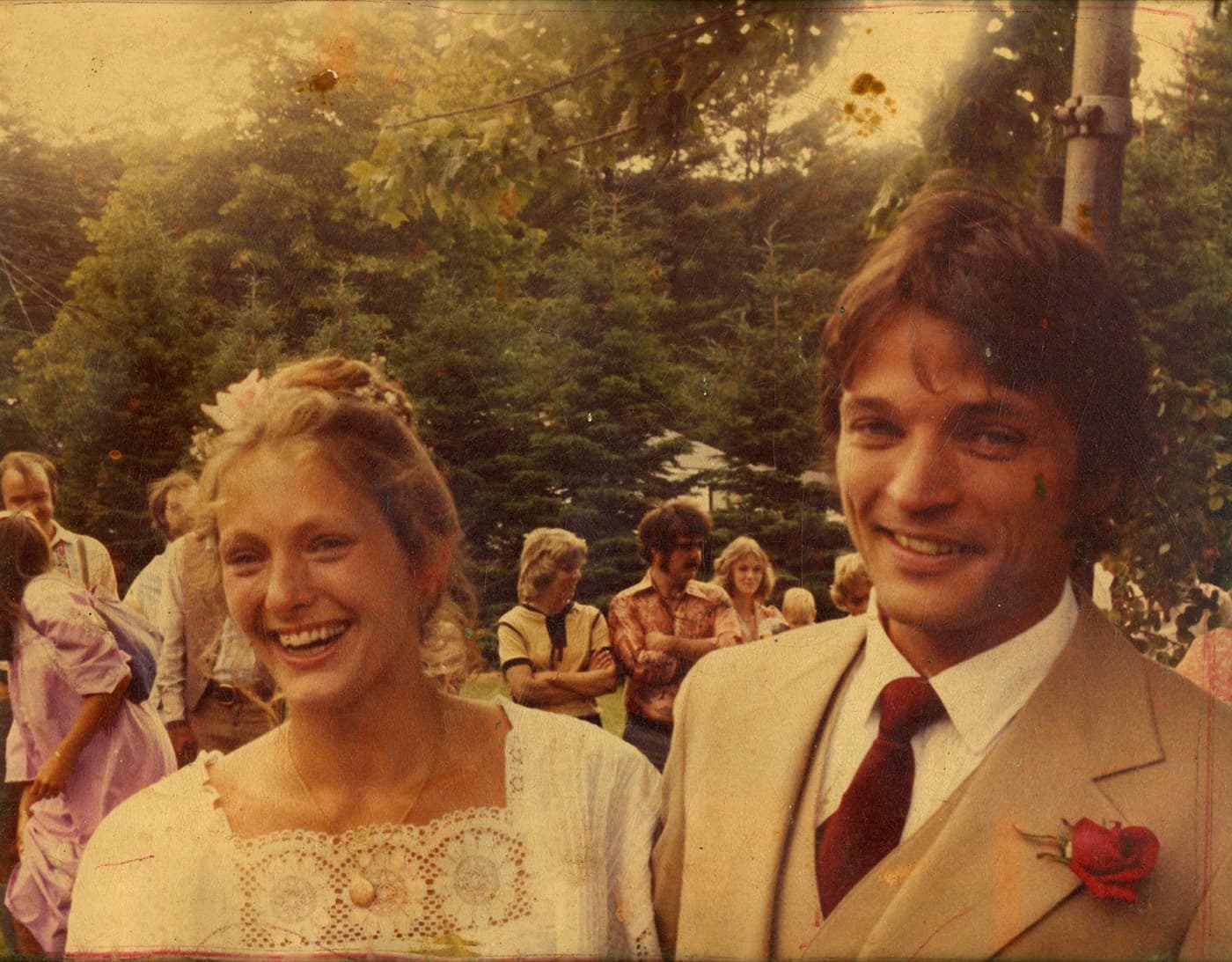 A warm faded photograph of Blake and Aletta laughing on their wedding day, spruce trees and guests behind them